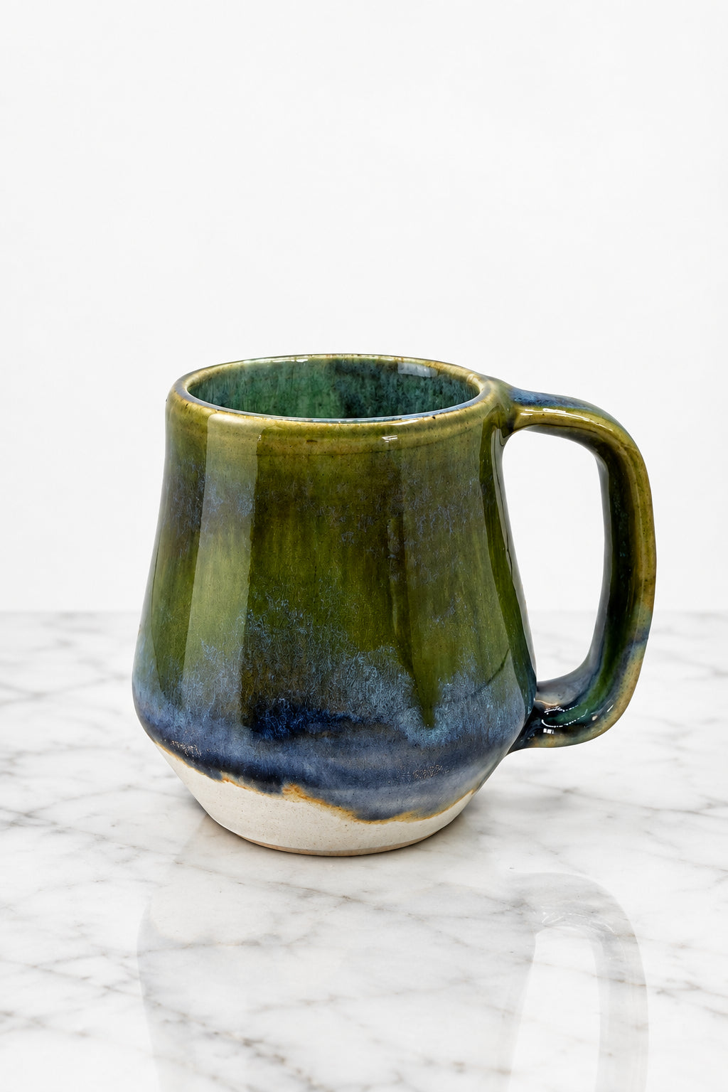 A handmade ceramic mug with a rustic tapered shape and a C-shaped handle, featuring a glossy glaze that transitions from olive green at the top to deep blue and dark brown at the base, with a white exposed clay rim.