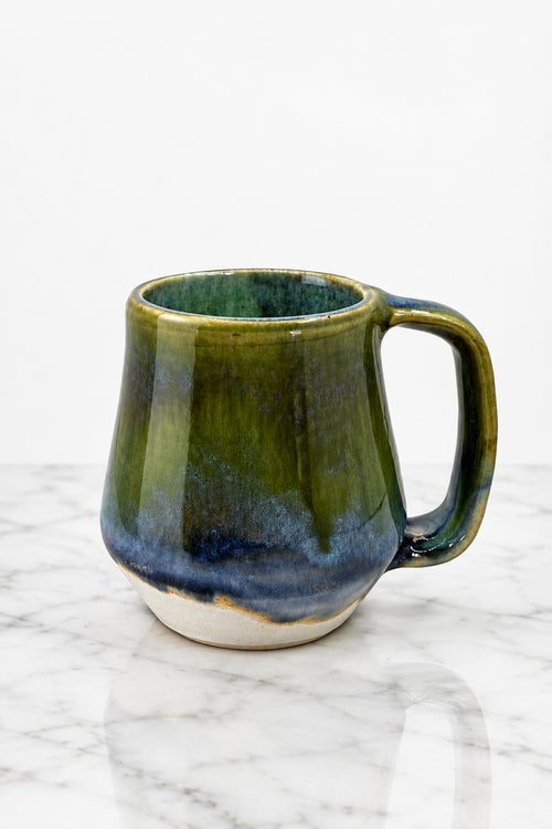 A handmade ceramic mug with a rustic tapered shape and a C-shaped handle, featuring a glossy glaze that transitions from olive green at the top to deep blue and dark brown at the base, with a white exposed clay rim.